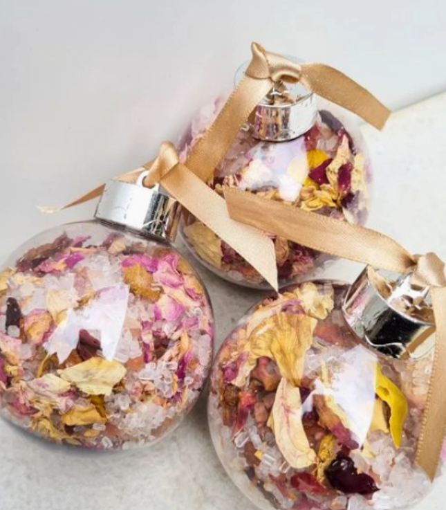 Three clear bath bomb christmas bauble ornaments filled with dried flowers, rose petals, and bath salt crystals, tied with beige ribbon and topped with silver caps.